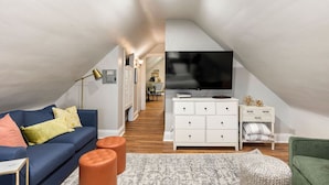Un appartement moderne de style « loft » avec un plafond en pente, un grand téléviseur à écran plat fixé au mur, un canapé bleu, une commode blanche et un fauteuil vert.
