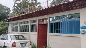 A building with barred windows, a red door, and a white car parked in front. - Se alquila casa humilde en Limón. (Limón)