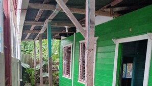 A covered outdoor area with green walls and a concrete floor, featuring a window with metal bars and a door leading to an interior space. - Se alquila casa humilde en Limón. (Limón)