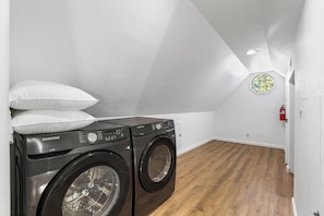 A laundry room with two Samsung washing machines, a white wall, wooden flooring, and a fire extinguisher on the wall.