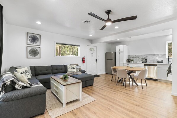 A modern living room with a sofa, coffee table, and dining area with chairs. There is a kitchen with cabinets and appliances.