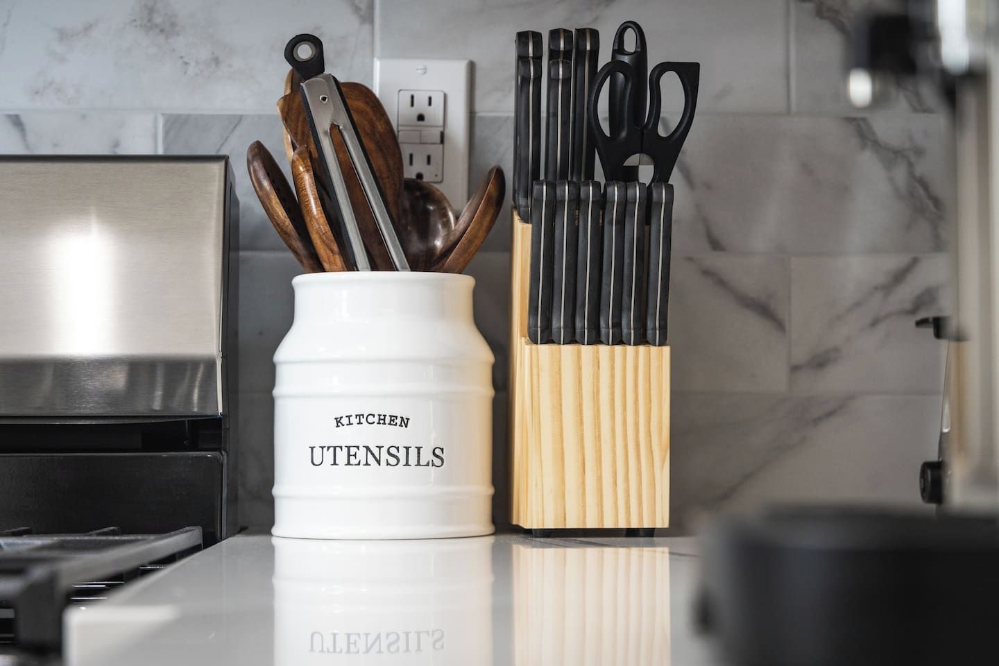 Eine Küchenarbeitsplatte mit einem weißen Behälter für Küchenutensilien mit der Aufschrift 'KITCHEN UTENSILS' und einem Holzblock für Küchenwerkzeuge.