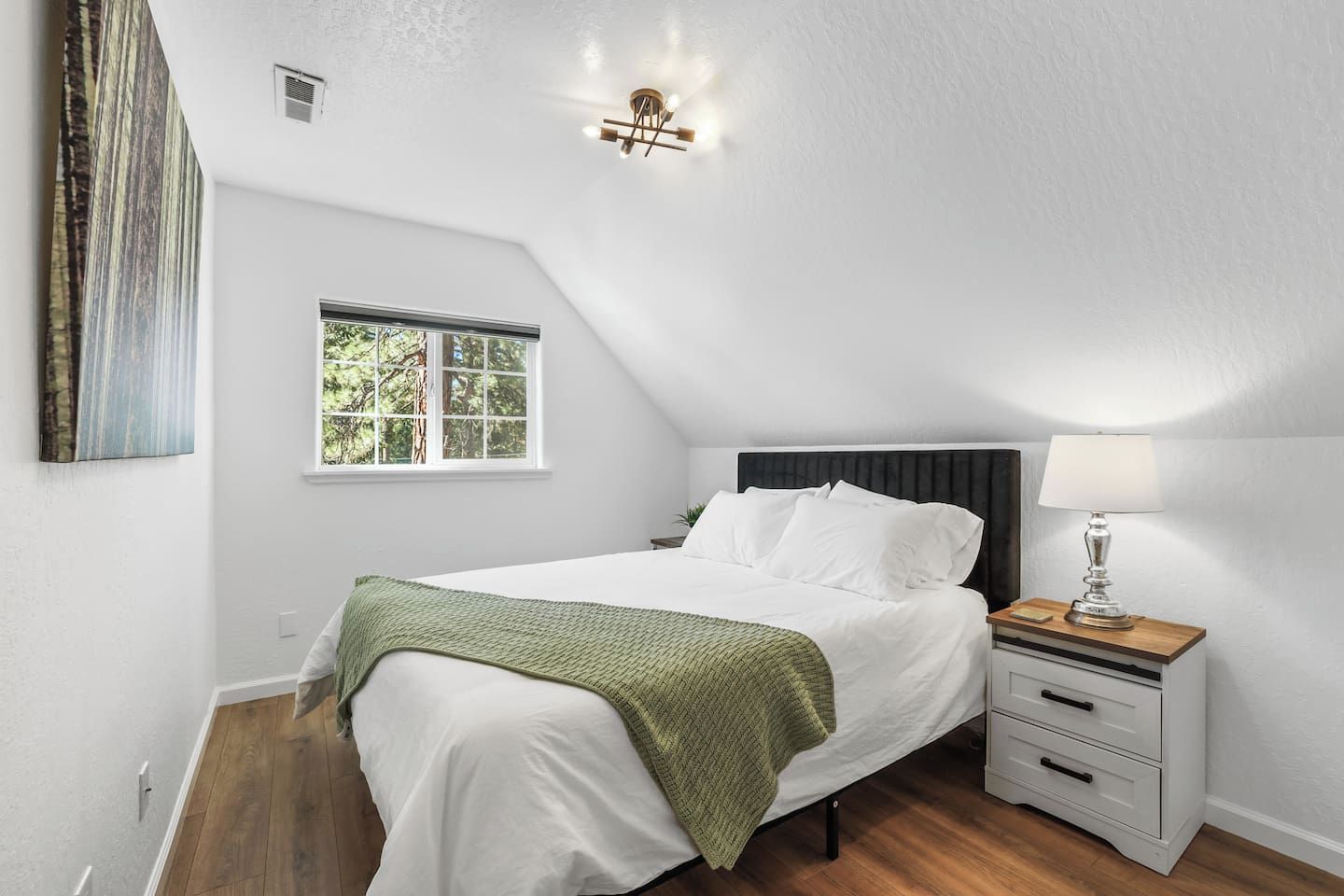 Un dormitorio con techo inclinado, cama con ropa blanca, mesita de noche con lámpara y ventana con vista a árboles.