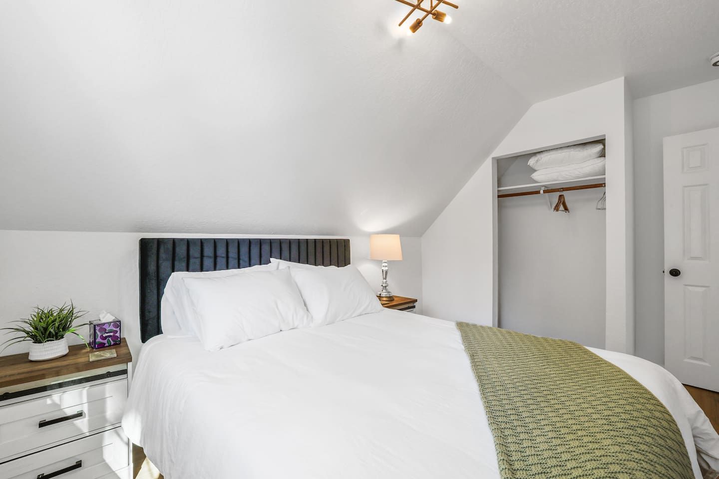A bedroom with a bed, bedside table, lamp, and a closet with hanging clothes.