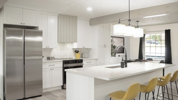 Una cocina moderna con muebles blancos, electrodomésticos de acero inoxidable, una isla central con taburetes y un amplio ventanal con vistas al exterior.