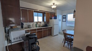 A compact kitchen with wooden cabinets, a white refrigerator, a coffee maker, and a dining area with a table and chairs. - Oceanfront Cottage (Charlo)