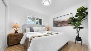 Ein Schlafzimmer mit einem großen Bett, Holz-Nachttischen, einem Fenster mit Blick, einer Pflanze und einem Gemälde an der Wand.