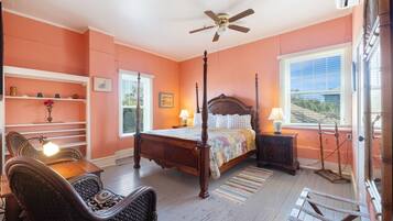 A bedroom with a four-poster bed, a ceiling fan, a window with blinds, a bedside table with a lamp, and a chair.