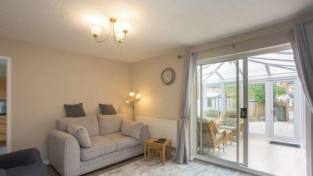 A cozy living room with a sofa, a small table, and a lamp. There is a large window with curtains and a view of the outside. - Sarah`s House: BBQ Parking 10mins To Alton Towers (Alton Towers & Staffordshire Villages)