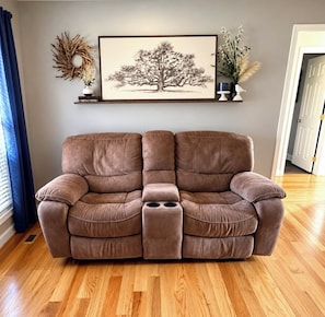 A brown sofa with a footrest in a room with wooden flooring, a framed tree artwork on the wall, and a door in the background.