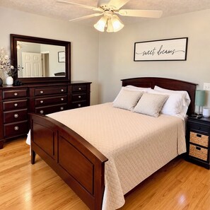 A bedroom with a wooden bed, a nightstand, a dresser, a ceiling fan, and a wall-mounted mirror.