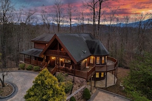 A log cabin with a wrap-around porch, multiple decks, and a view of the surrounding forest and mountains.