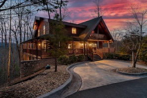 A log cabin with a stone foundation, a wrap-around porch, and a curved driveway.
