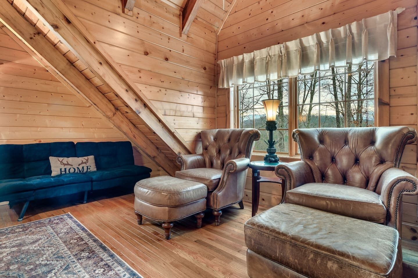 A cozy room with a sloped ceiling, a dark blue sofa, leather armchairs, a matching ottoman, and a window with a view of trees.