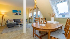 A cozy living room with a grey sofa, a wooden dining table with chairs, and a large window with a view of the sky. - Tom - 3-room vacation apartment on the 1st floor in Westerland with terrace. (Sylt)