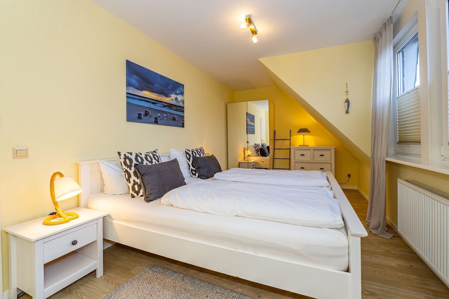 Ein Schlafzimmer mit einem großen Bett, einem Nachttisch, einer Lampe, einem Gemälde, einem Spiegel, einer Leiter und einem Fenster mit Vorhängen.