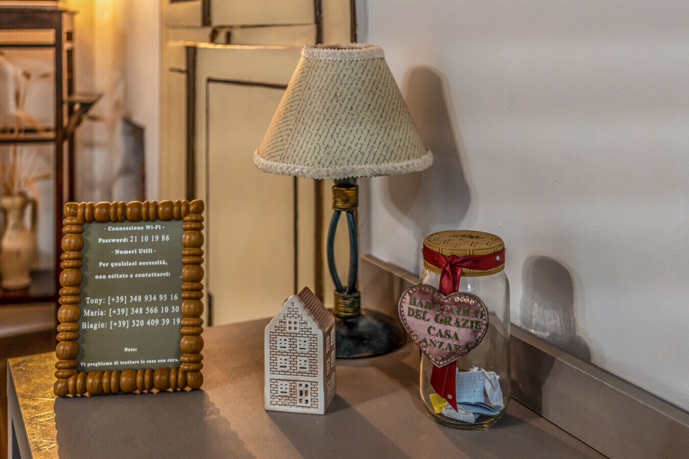 Una lámpara, un cartel enmarcado con información de contacto, una pequeña maqueta de una casa y una botella con forma de corazón y una etiqueta.
