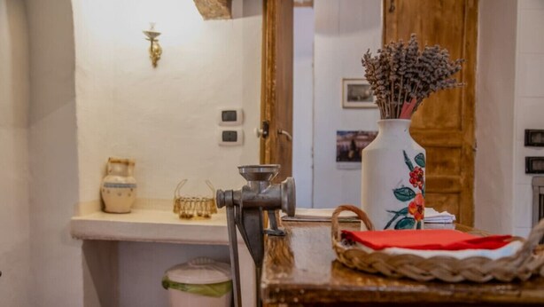 A rustic kitchen with a wooden table, a vintage meat grinder, a white ceramic pitcher, and a vase with dried flowers.
