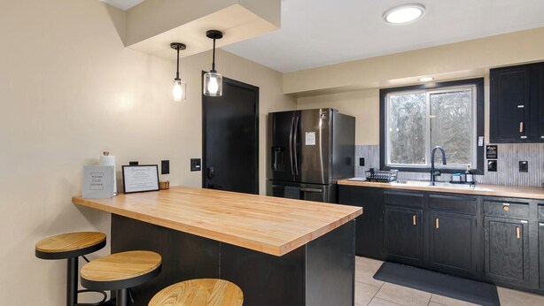 A modern kitchen with a central island, bar stools, stainless steel appliances, and black cabinetry. - Zen Den Oasis: Retreat in Nature, 4-Bedroom Home on 10 Acres of Private Land (Cottage Grove)