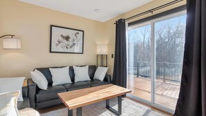 A modern living room with a black leather sofa, a wooden coffee table, and a large window with a view of a wooden deck. - Zen Den Oasis: Retreat in Nature, 4-Bedroom Home on 10 Acres of Private Land (Cottage Grove)