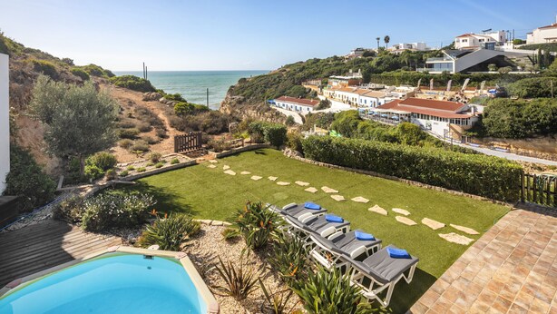 Zona de piscina con vista al mar y jardín con sillones de descanso.