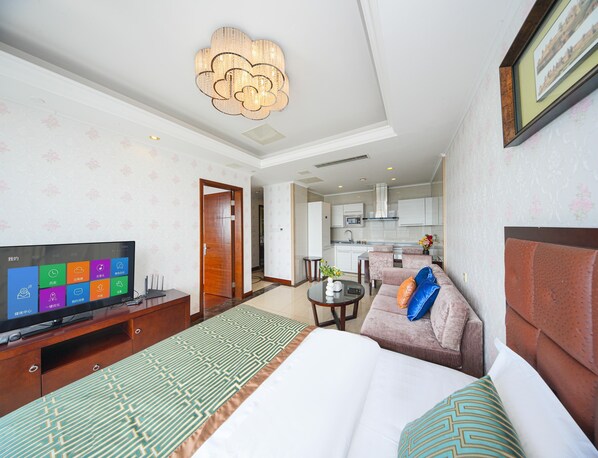 Habitación de hotel con cama, sofá, televisión, mesa de comedor y cocina al fondo.