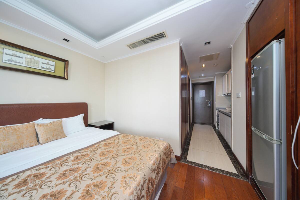 Une chambre d’hôtel comprenant un lit, une table de chevet, un réfrigérateur, un coin cuisine et un couloir menant à une autre pièce.