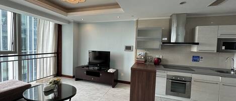 A modern living room with a sofa, a glass coffee table, and a TV. There is a kitchen area with a microwave, stove, and cabinets. The room has large windows and a chandelier.