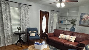 A living room with a brown leather sofa, a blue armchair, a wicker ottoman, and a patterned rug. - Gorgeous quiet and cozy 3 bedroom/ 2 bathroom home awaits you! (Uvalde)