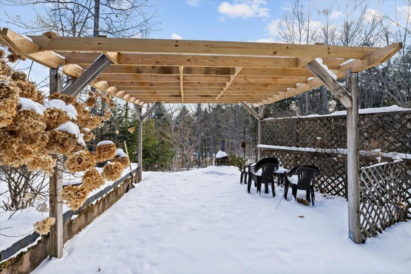 Un espacio al aire libre cubierto con una pérgola de madera, suelo cubierto de nieve y un conjunto de sillas.