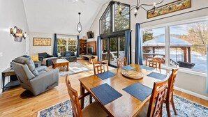 Ein geräumiges Wohnzimmer mit Kamin, einem Essbereich mit großem Holztisch und Blick auf eine verschneite Landschaft durch große Fenster.