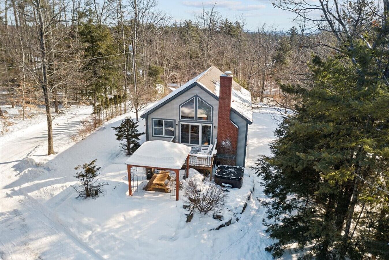 Une cabane dotée d’une grande fenêtre, d’une cheminée et d’une terrasse recouverte de neige.