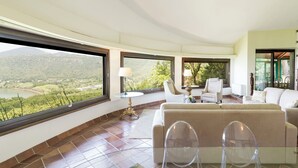 A spacious living room with a large window offering a scenic view of a lake and mountains. There is a beige sofa and two clear chairs.