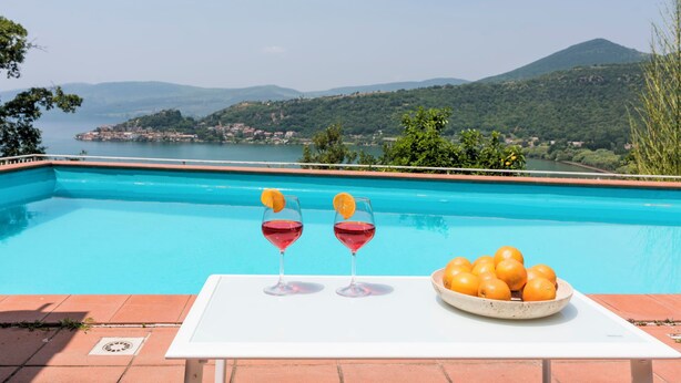 A poolside table with two glasses of red drink and a bowl of oranges, overlooking a scenic view of a lake and mountains.