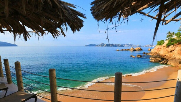 A beach view with a rope barrier, lounge chairs, and a thatched roof structure. - Suite 1808 by Suites Victoria Acapulco (Acapulco de Juárez)