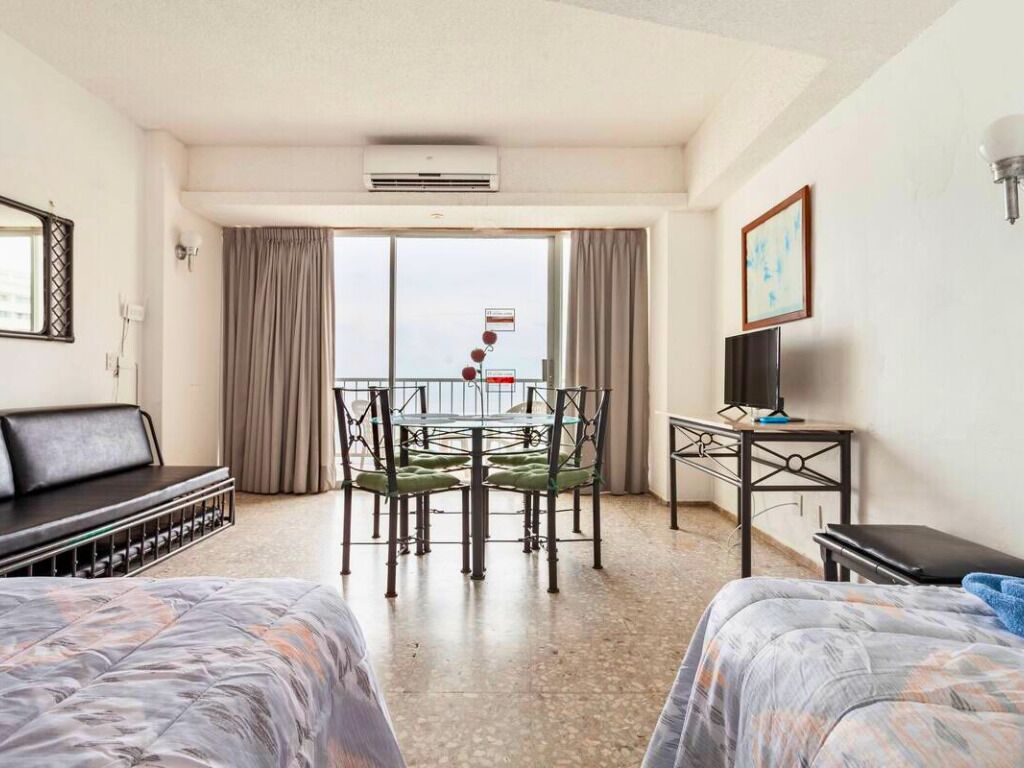 A hotel room with two beds, a dining table with chairs, a TV, and a balcony with a view.