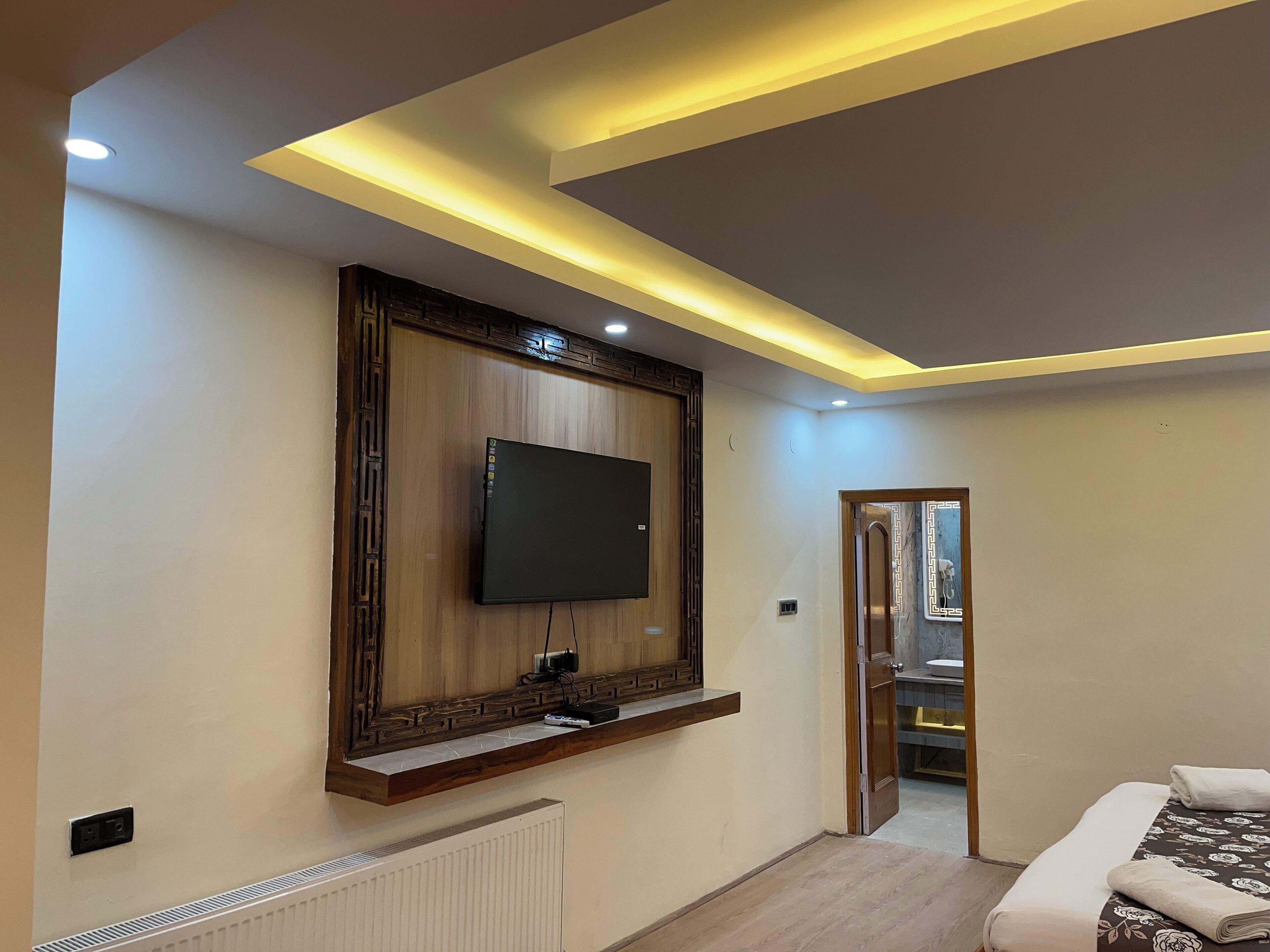 A modern bedroom with a mounted TV, wooden paneling, a built-in shelf, a radiator, and a door leading to another room.