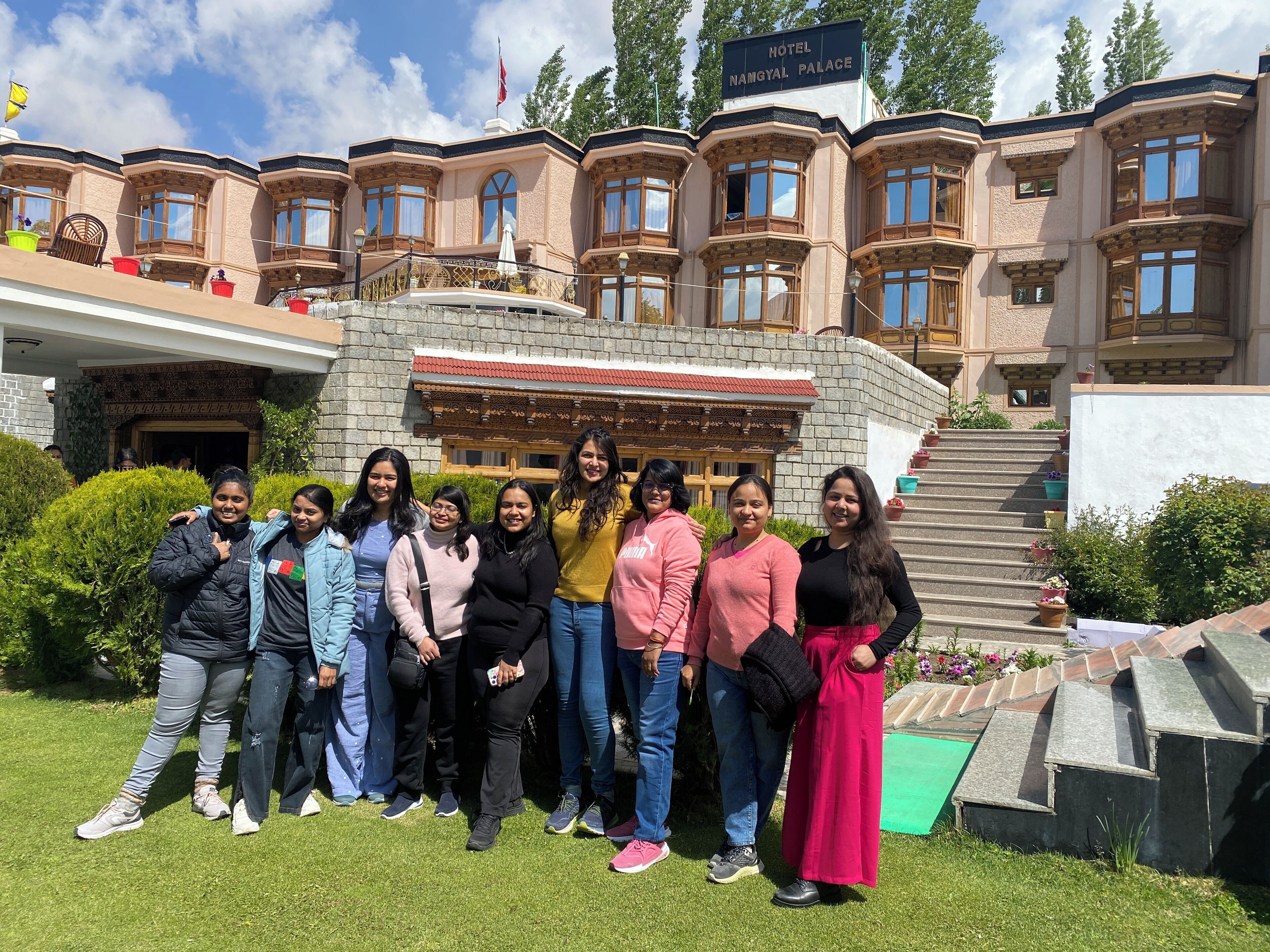 A group of people standing in front of a building labeled "Hotel Namgyal Palace" with a stone facade, multiple windows, and a red-tiled roof.