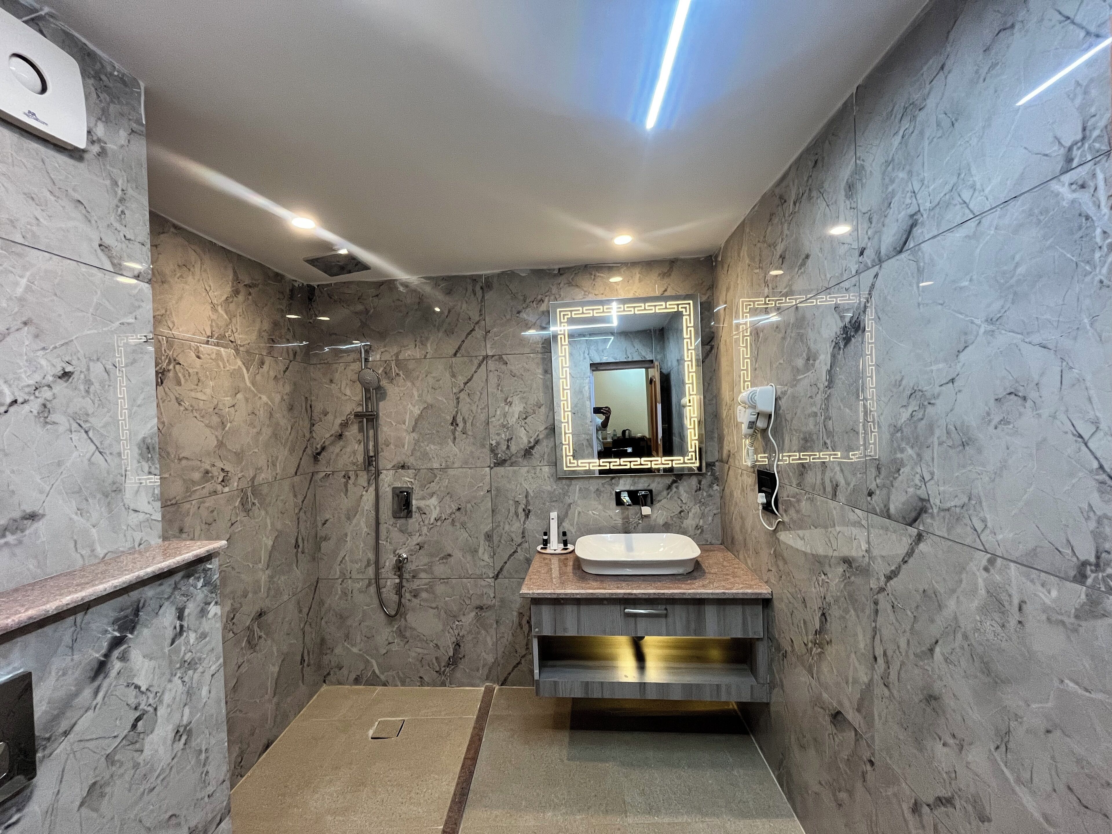 Un baño moderno con pared de mármol, tocador con lavabo, espejo con iluminación y zona de ducha.