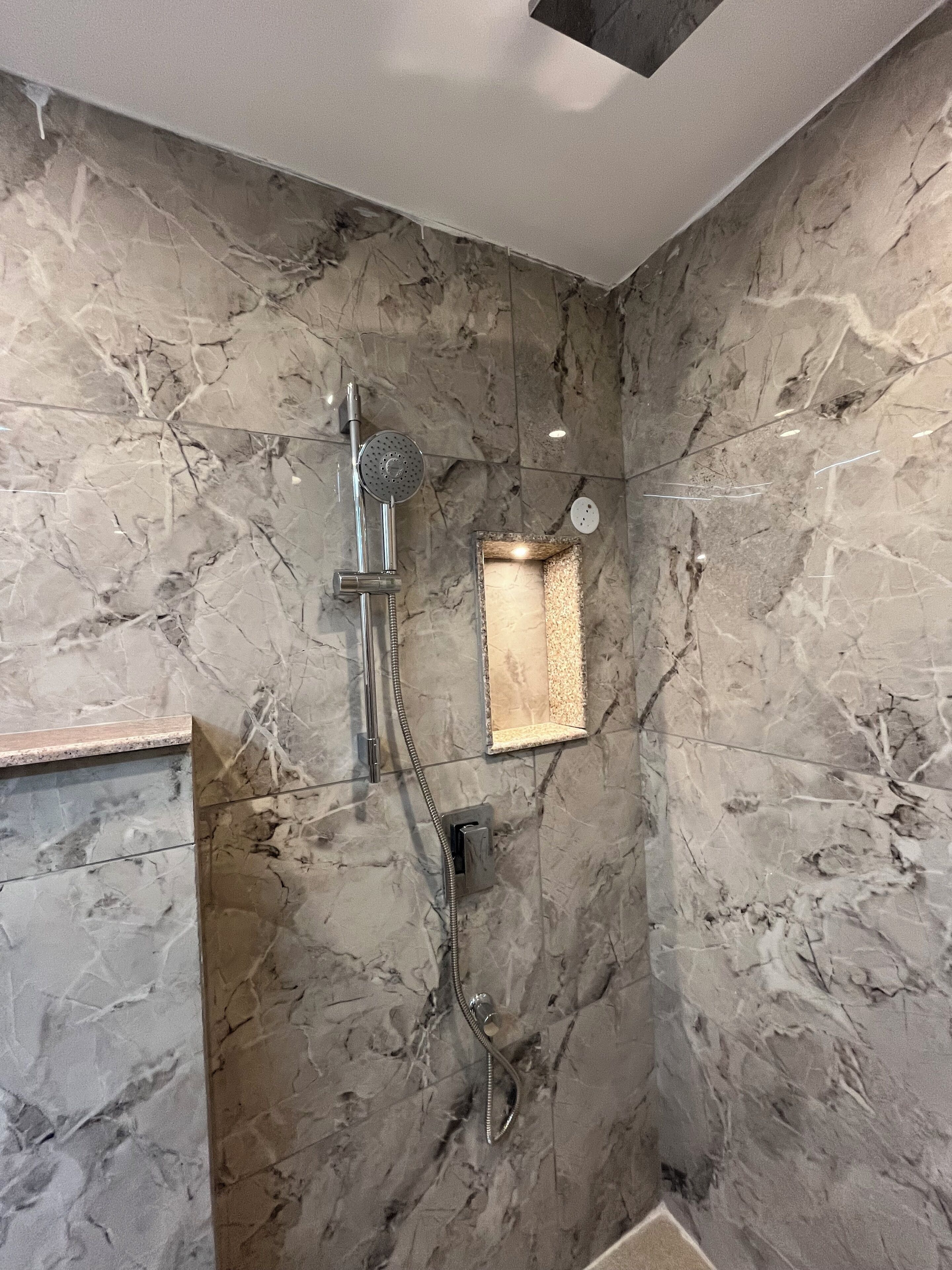 Une salle de bain avec un mur en marbre, une douche équipée d’un pommeau de douche à main et un luminaire fixé au mur.