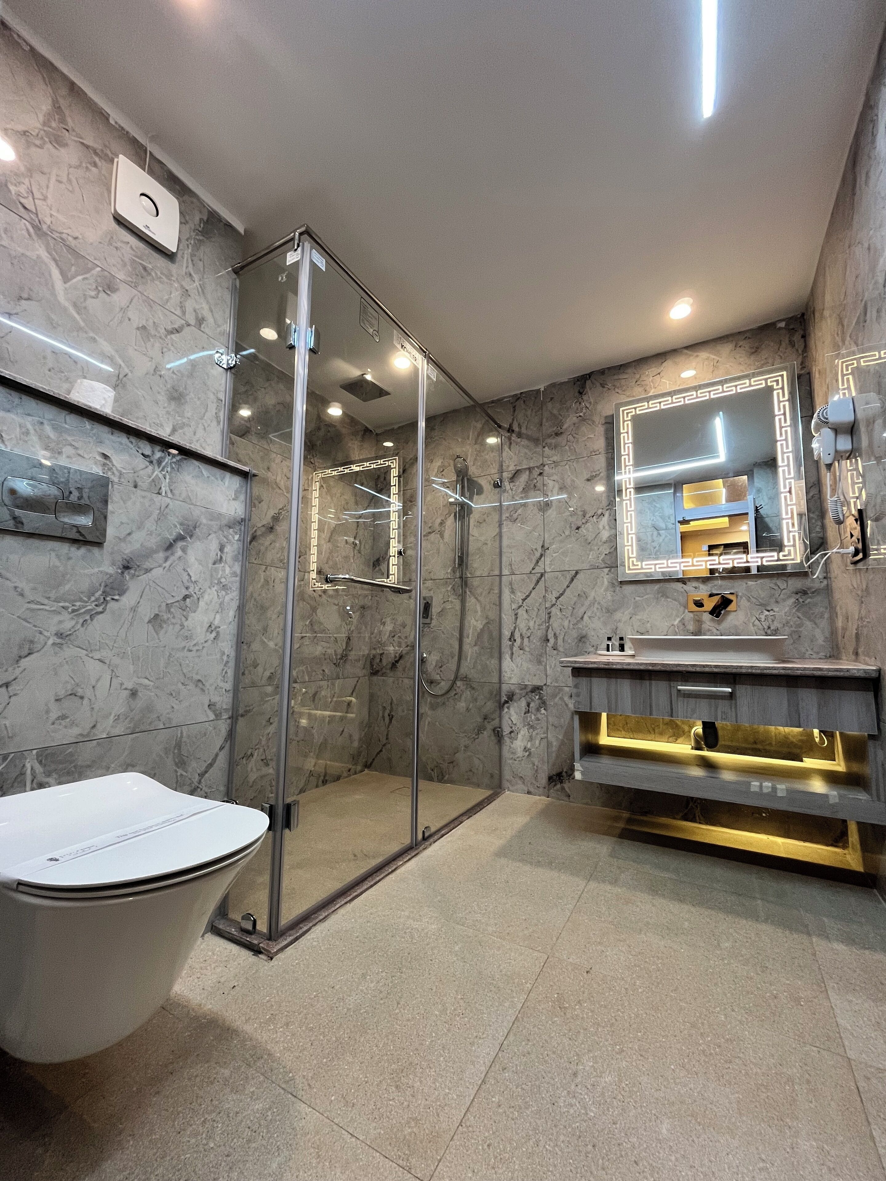 Een moderne badkamer met een douchecabine van glas, een betegelde wand van marmer, een wastafel met gouden accenten en een grote spiegel.
