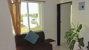 A living room with a brown sofa, a blue pillow, a potted plant, and a window with curtains. - San Cristobal Surf House (Puerto Baquerizo Moreno)