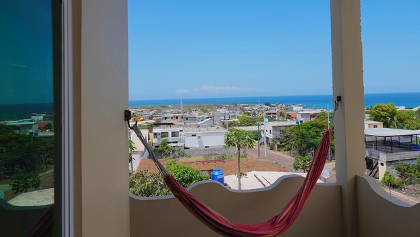 A view from a balcony with a hammock, overlooking a coastal town with buildings and the sea. - San Cristobal Surf House (Puerto Baquerizo Moreno)