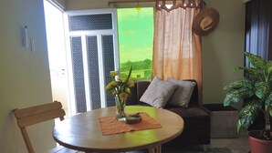 A cozy living area with a round wooden table, a sofa with cushions, a potted plant, and a view through a window with a curtain. - San Cristobal Surf House (Puerto Baquerizo Moreno)