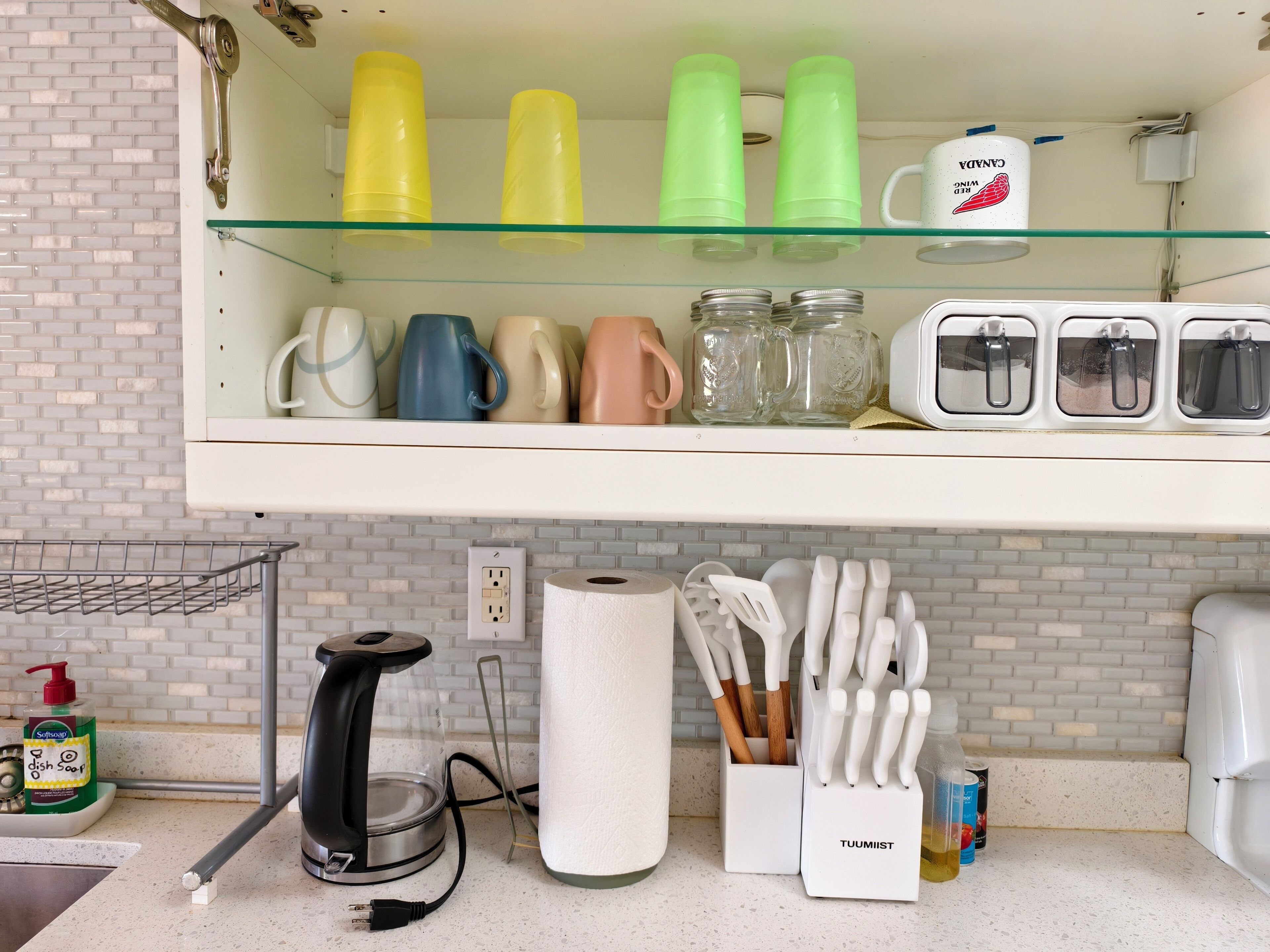 A kitchen with a white countertop, a dish soap dispenser, a kettle, a paper towel holder, and a knife block.