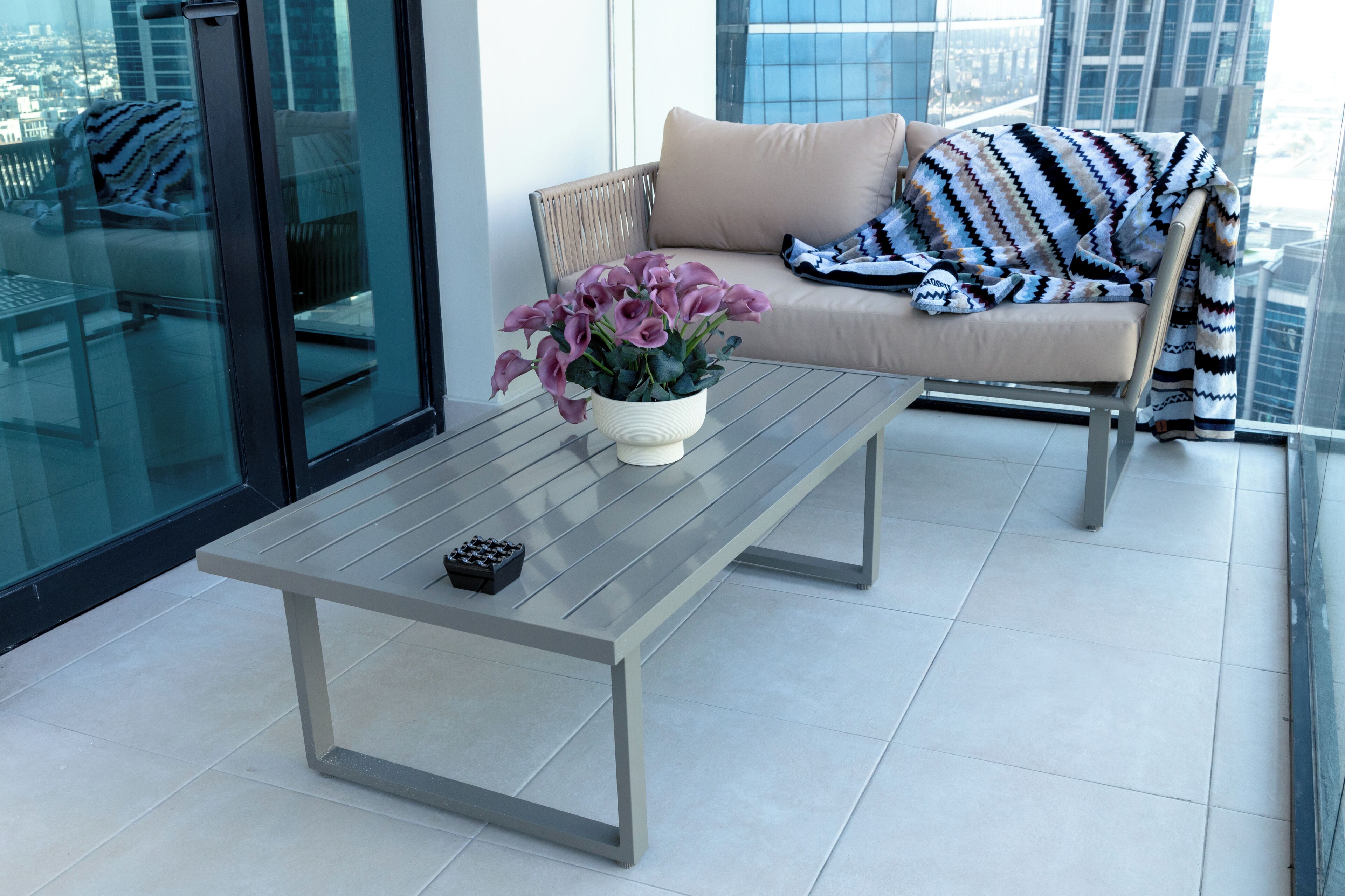 A modern balcony with a glass railing, a beige sofa with patterned throw, and a potted plant with purple flowers.