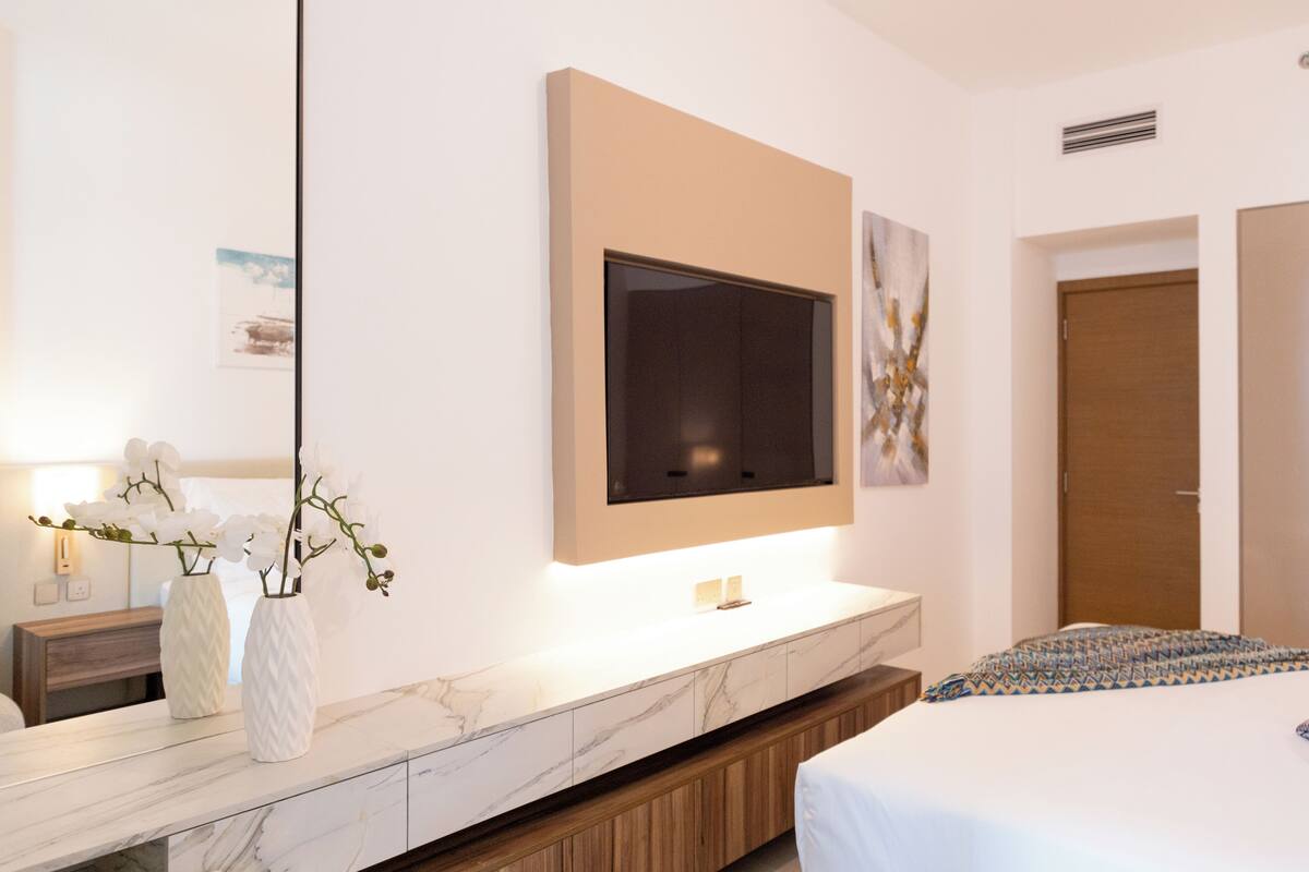 Ett modernt hotellrum med en stor säng, en tv som är monterad på väggen, ett sängbord i marmor och en träbänk.
