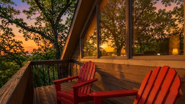 A wooden deck with two red Adirondack chairs, a railing, and a view of trees and a sunset through large windows. - 3 bedroom 3 bath, expansive views Treehouse feel (Galena)