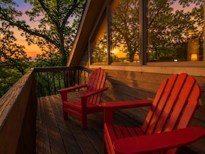 Een houten terras met twee rode Adirondack stoelen, een leuning en uitzicht op bomen en een zonsondergang door grote ramen.
