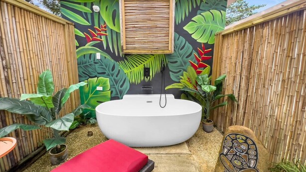 A spa area with a large oval bathtub, a red massage table, and a wall mural with tropical leaves and flowers. - Casa Luquillo | Cozy Retreat House W/ BBQs & Tub (Luquillo)